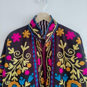 May include: A dark brown jacket with colorful floral embroidery. The jacket features bright pink, yellow, blue, and orange floral designs. The collar and edges are also decorated with the same vibrant colors. The jacket is hanging on a wooden hanger.