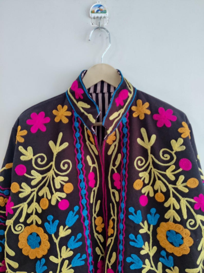 May include: A dark brown jacket with colorful floral embroidery. The jacket features bright pink, yellow, blue, and orange floral designs. The collar and edges are also decorated with the same vibrant colors. The jacket is hanging on a wooden hanger.