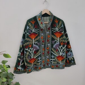 May include: A dark green velvet jacket with colorful floral embroidery. The jacket features orange, red, blue, and white flower designs. It has a collared neckline and is hanging on a wooden hanger.