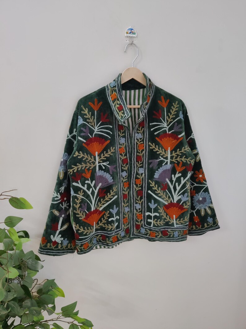 May include: A dark green velvet jacket with colorful floral embroidery. The jacket features orange, red, blue, and white flower designs. It has a collared neckline and is hanging on a wooden hanger.