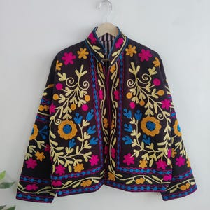 May include: A dark brown jacket with vibrant floral embroidery in yellow, pink, and blue. The jacket features a high collar and is adorned with intricate embroidered patterns. The design includes a mix of floral and leaf motifs, creating a colorful and detailed garment.