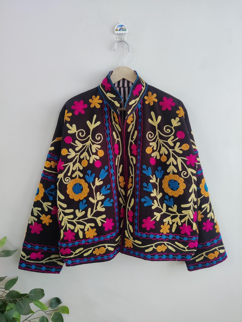 May include: A dark brown jacket with vibrant floral embroidery in yellow, pink, and blue. The jacket features a high collar and is adorned with intricate embroidered patterns. The design includes a mix of floral and leaf motifs, creating a colorful and detailed garment.