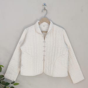 Puede incluir: Chaqueta acolchada de color crema con cuello mandarín y mangas largas. La chaqueta presenta una serie de pequeños cierres decorativos en la parte delantera. Cuelga de una percha de madera sobre un fondo neutro. Una elegante adición a cualquier armario.