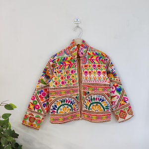 Peut inclure: Veste brodée colorée avec un col montant. La veste présente un motif patchwork vibrant avec des motifs floraux et géométriques dans les tons de rose, jaune, vert et bleu. La veste est suspendue à un mur blanc.