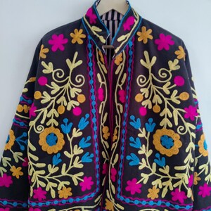 May include: A dark brown jacket with colorful floral embroidery. The jacket features yellow, pink, blue, and orange embroidered flowers and vines. The collar and edges are trimmed with a contrasting fabric. The jacket is hanging on a silver hanger.