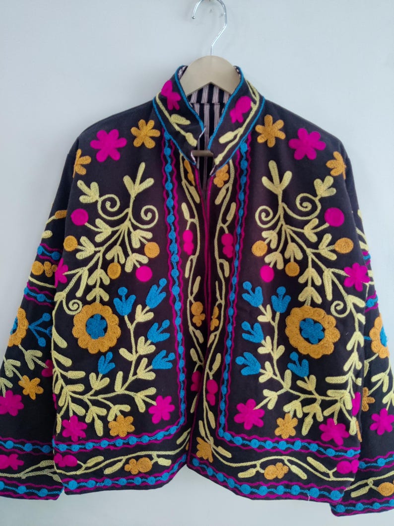 May include: A dark brown jacket with colorful floral embroidery. The jacket features yellow, pink, blue, and orange embroidered flowers and vines. The collar and edges are trimmed with a contrasting fabric. The jacket is hanging on a silver hanger.