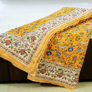 May include: A yellow floral quilt with a white and floral border is folded on a bed. The quilt features a repeating pattern of red, blue, and white flowers on a yellow background. The border has a striped yellow pattern.