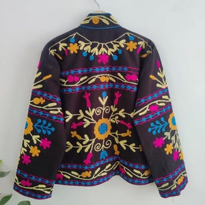 May include: A dark brown jacket with colorful floral embroidery. The embroidery features yellow, blue, pink, and orange flowers and patterns. The jacket is hanging on a silver hook against a white wall.