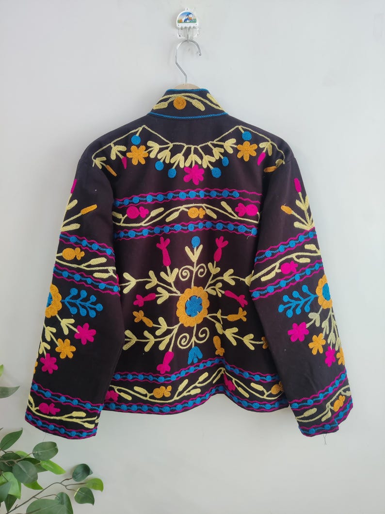May include: A dark brown jacket with colorful floral embroidery. The embroidery features yellow, blue, pink, and orange flowers and patterns. The jacket is hanging on a silver hook against a white wall.