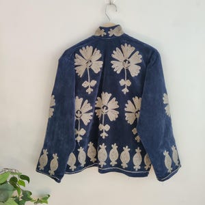 May include: A navy blue jacket with intricate beige floral embroidery. The jacket features a high collar and long sleeves. The floral design is repeated across the back and sleeves, creating a cohesive and detailed pattern. The jacket is hanging on a wooden hanger.