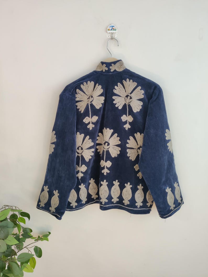 May include: A navy blue jacket with intricate beige floral embroidery. The jacket features a high collar and long sleeves. The floral design is repeated across the back and sleeves, creating a cohesive and detailed pattern. The jacket is hanging on a wooden hanger.
