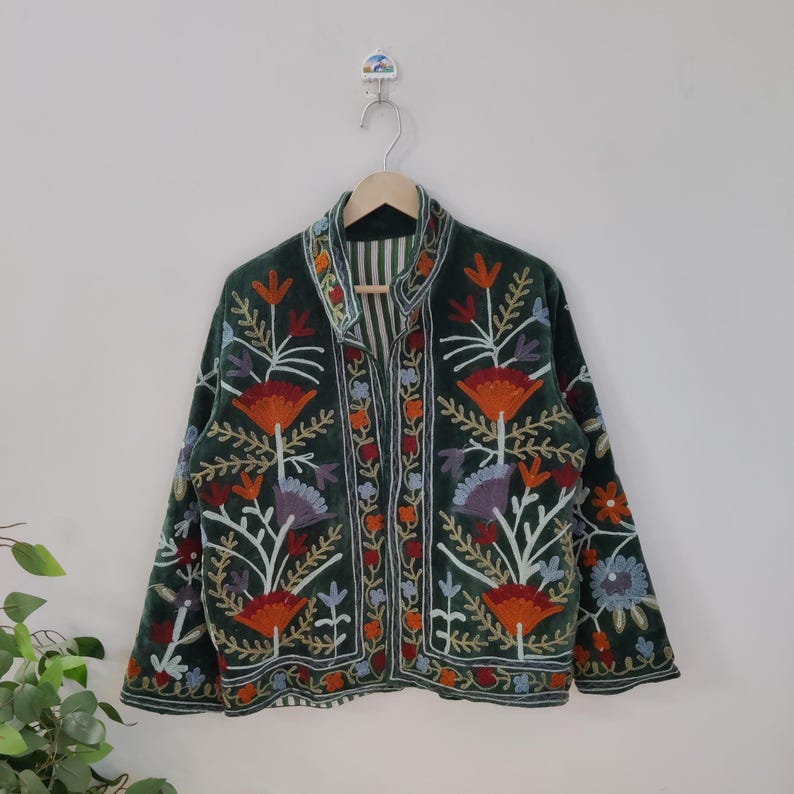 May include: A dark green velvet jacket with colorful floral embroidery. The jacket features a collared neckline and a button-up front. The embroidery includes orange, red, purple, and blue flowers with green leaves. The jacket is hanging on a wooden hanger.