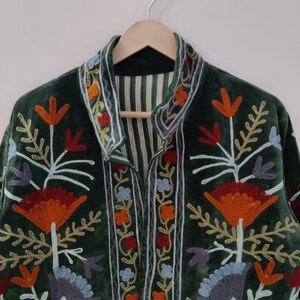 May include: A dark green velvet jacket with colorful floral embroidery. The jacket features orange, red, blue, and yellow flower designs with white stems and leaves. The collar and edges are trimmed with embroidered details. The jacket is hanging on a wooden hanger.