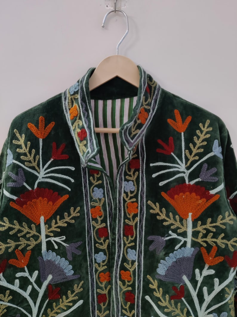 May include: A dark green velvet jacket with colorful floral embroidery. The jacket features orange, red, blue, and yellow flower designs with white stems and leaves. The collar and edges are trimmed with embroidered details. The jacket is hanging on a wooden hanger.