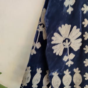 May include: Close-up of a section of a dark blue velvet garment with intricate white floral embroidery. The embroidery features large flowers and smaller, stylized floral motifs. The fabric has a soft, textured appearance.