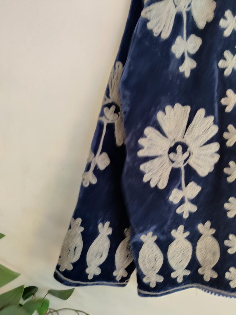 May include: Close-up of a section of a dark blue velvet garment with intricate white floral embroidery. The embroidery features large flowers and smaller, stylized floral motifs. The fabric has a soft, textured appearance.