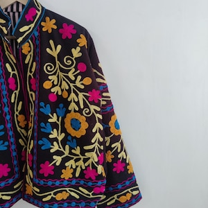 May include: A dark brown jacket with colorful floral embroidery. The jacket features yellow, pink, blue, and orange flowers and vines. The collar and cuffs are trimmed with a contrasting color. The jacket is hanging on a wooden hanger.
