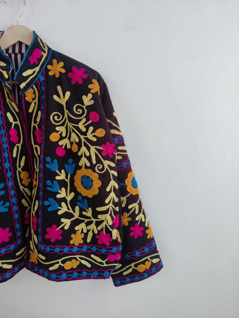 May include: A dark brown jacket with colorful floral embroidery. The jacket features yellow, pink, blue, and orange flowers and vines. The collar and cuffs are trimmed with a contrasting color. The jacket is hanging on a wooden hanger.