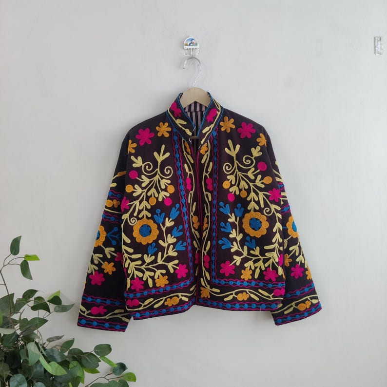 May include: A dark brown jacket with colorful floral embroidery. The jacket features yellow, blue, and pink flowers and vines. The jacket is hanging on a white wall.