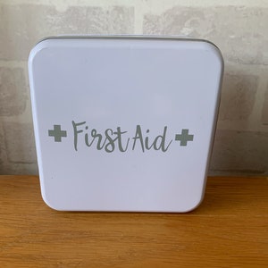 May include: White metal first aid kit with the words "First Aid" in grey lettering and a plus sign on either side of the text.