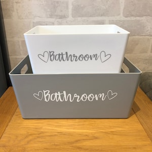 May include: Two grey plastic storage bins with white lids. The bins are stacked on top of each other. The front of each bin has the word "Bathroom" written in white with a heart on either side of the word.