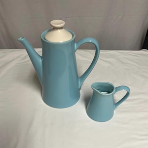 Könnte beinhalten: Eine hellblaue Keramik-Kaffeekanne mit weißem Deckel und ein passender Sahnekännchen. Die Kaffeekanne hat einen langen Ausguss und einen gebogenen Griff. Das Sahnekännchen hat einen kleinen Griff. Beide Teile stehen auf einer weißen Oberfläche.