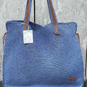 May include: A blue crocheted tote bag with brown leather handles. The bag has a small tag that says "Handmade with Love".