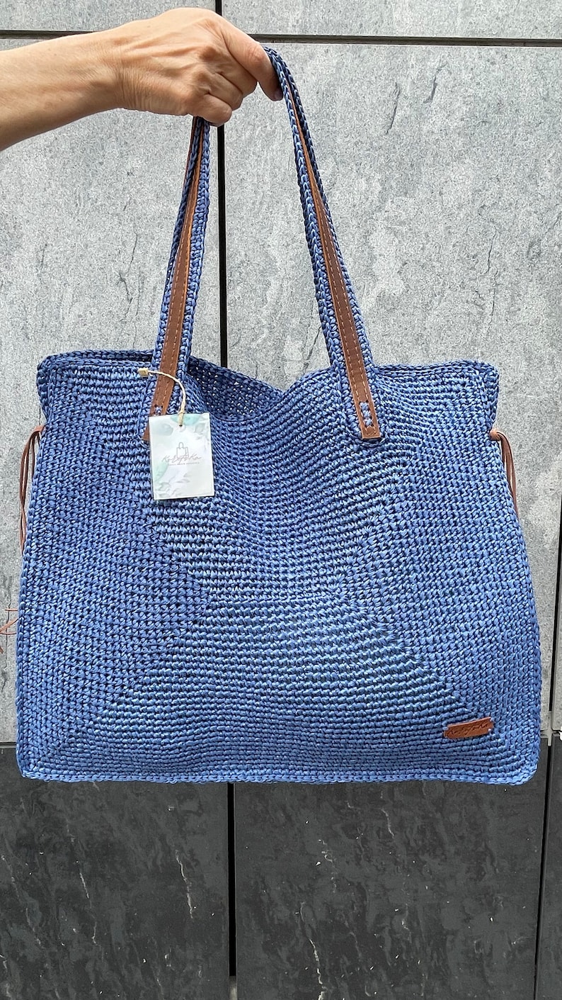 May include: A blue crocheted tote bag with brown leather handles. The bag has a small tag that says "Handmade with Love".