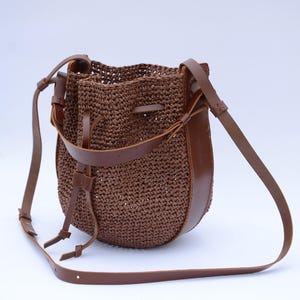 May include: A brown woven bucket bag with leather accents and a long shoulder strap. The bag features a drawstring closure and a rounded shape. The leather details are a darker shade of brown, adding contrast to the woven body.