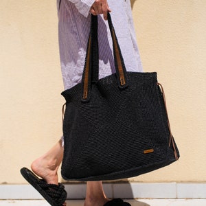 May include: A black crocheted tote bag with brown leather handles and a brown leather tag. The bag is being held by a person wearing a striped shirt and black sandals.