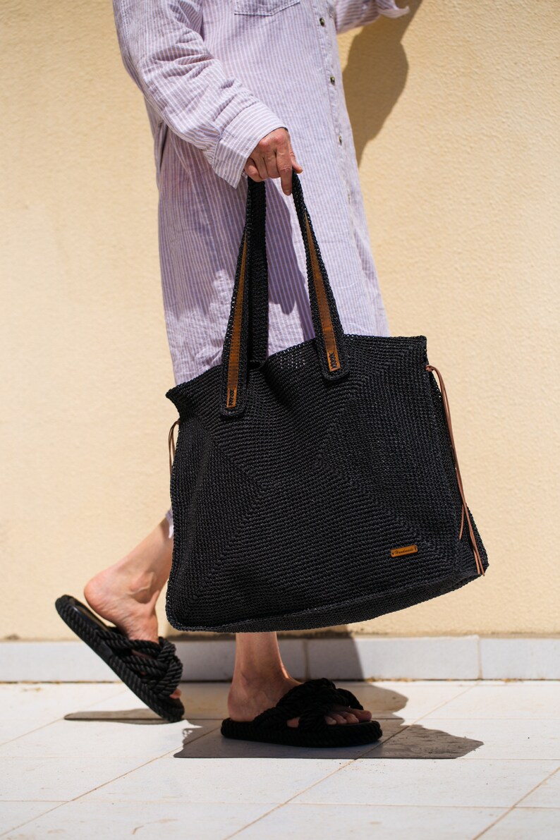 May include: A black crocheted tote bag with brown leather handles and a brown leather tag. The bag is being held by a person wearing a striped shirt and black sandals.