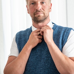 Op de afbeelding: Een man past de kraag van een blauwe gebreide vest aan. Hij draagt er een wit overhemd onder. De vest heeft een textuur met spikkels.