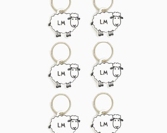 May include: Six white sheep keychains with black eyes, noses, and legs. Each keychain has a silver ring and the letters "LM" printed on the sheep's body.