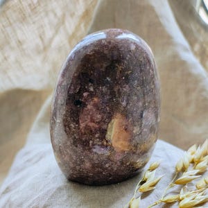 Puede incluir: Una piedra preciosa lisa y pulida, de color púrpura y rosa, con una forma natural y orgánica. La piedra descansa sobre un fondo de tela marrón claro.