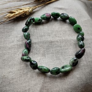 Puede incluir: Un brazalete de cuentas con piedras preciosas ovaladas verdes y rojas. El brazalete se muestra sobre una tela de tono neutro, con espigas de trigo en el fondo. Las piedras tienen un acabado pulido.