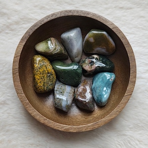 Peut inclure: Un ensemble de huit pierres naturelles polies, multicolores, dans un bol en bois. Les pierres sont de différentes nuances de vert, bleu, marron et jaune.
