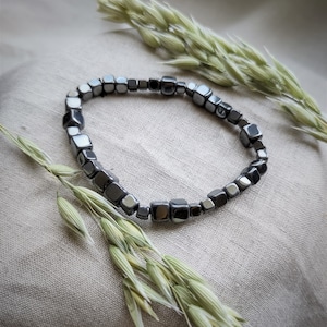 Könnte beinhalten: Ein schwarzes Hämatit-Armband mit quadratischen Perlen. Das Armband liegt auf einem hellbeigen Stoffhintergrund mit getrockneten Haferhalmen.