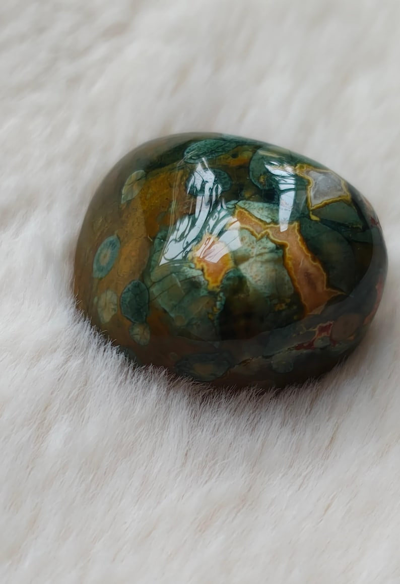 May include: A smooth, polished green, brown, and orange jasper stone with a unique pattern. The stone is oval shaped and has a glossy finish.