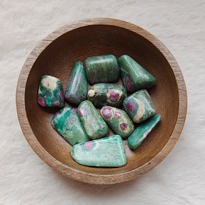 Può includere: Una ciotola di legno piena di pietre rotonde verdi e rosa. Le pietre hanno un aspetto maculato con una miscela di colori verdi e rosa.