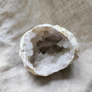 Op de afbeelding: Een geode met een ruwe, beige buitenkant en een holte gevuld met sprankelende, witte kristalformaties. De geode is open en onthult de binnenste kristallen. De achtergrond is een neutrale, getextureerde stof.