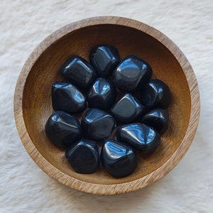 Può includere: Una ciotola di legno piena di pietre di ossidiana nera rotonde. Le pietre sono lisce e levigate, con un bagliore scuro e vetroso.