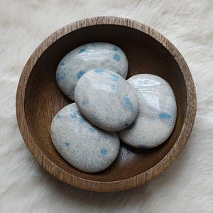 Könnte beinhalten: Vier glatte, weiße Steine mit blauen Flecken sind in einer Holzschale angeordnet. Die Steine sind hellgrau mit kleinen, verstreuten blauen Flecken.