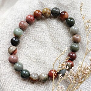 May include: A bracelet made of round, earth-toned gemstones in shades of red, brown, green, and grey. The beads are strung together to form a circular shape. The bracelet is displayed on a neutral linen surface.