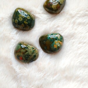 May include: Four green, brown, and white ocean jasper stones on a white fuzzy surface.