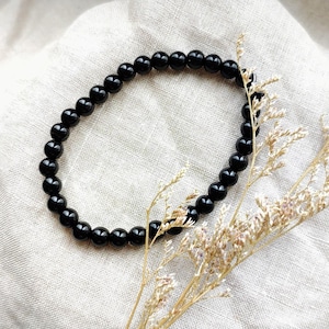 May include: A black beaded bracelet made of small, round beads. The bracelet is presented on a neutral linen cloth, with dried floral accents. The beads appear to be approximately 0.6 cm in diameter.