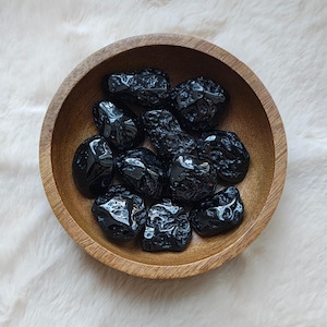 Könnte beinhalten: Eine Schale mit schwarzen, getrommelten Obsidiangesteinen. Die Steine sind glatt und haben eine glänzende, glasartige Oberfläche.