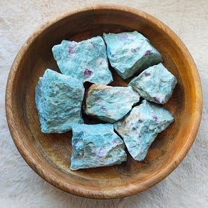 Op de afbeelding: Een verzameling van zes ruwe, tealgroene en roze robijn-zoisiet kristallen in een houten kom. De kristallen zijn van verschillende groottes en vormen, sommige vertonen een meer uitgesproken roze kleur.