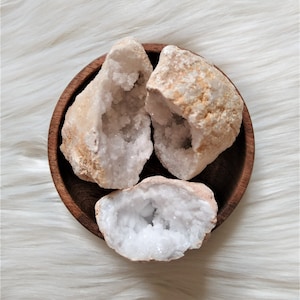 Peut inclure: Trois géodes aux intérieurs cristallins blancs et aux extérieurs beiges sont présentées dans un bol en bois foncé. Les géodes sont ouvertes, révélant les formations cristallines scintillantes. Le bol repose sur une surface blanche et duveteuse.