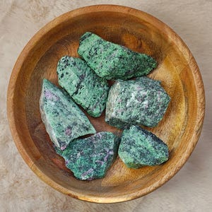 Op de afbeelding: Een houten kom gevuld met zes ruwe zultanite edelstenen in groen en roze.