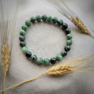 Peut inclure: Un bracelet de pierres précieuses vert et noir avec des perles rondes. Le bracelet est posé sur un fond de tissu brun clair.